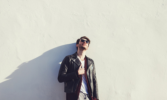 Trendy guy with leather jacket and eyeglasses standing by wall. copyspace