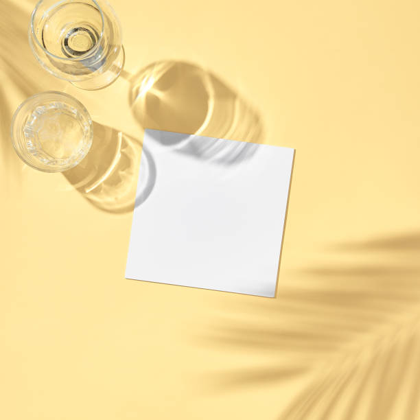 istock Glass with white paper card flat lay. Pastel yellow table in sunlight with palm leaf shadow. Wedding, birthday, tropical summer day concept 1302793326