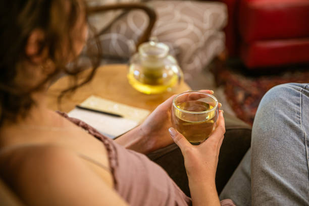 Young woman drinking loose leaf camomile tea Over the shoulder shot of a young drinking loose leaf camomile tea. She is sitting on a brown leather occasional chair in the living room with the glass teapot on the small wooden table beside her. glass cup of boiling water stock pictures, royalty-free photos & images