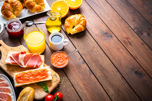Spanish tomato and ham toast, croissant, coffee, orange juice. Traditional Mediterranean breakfast or lunch on wooden table. The composition is at the left of an horizontal frame leaving useful copy space for text and/or logo at the right. High angle view. High resolution 42Mp studio digital capture taken with SONY A7rII and Zeiss Batis 40mm F2.0 CF lens