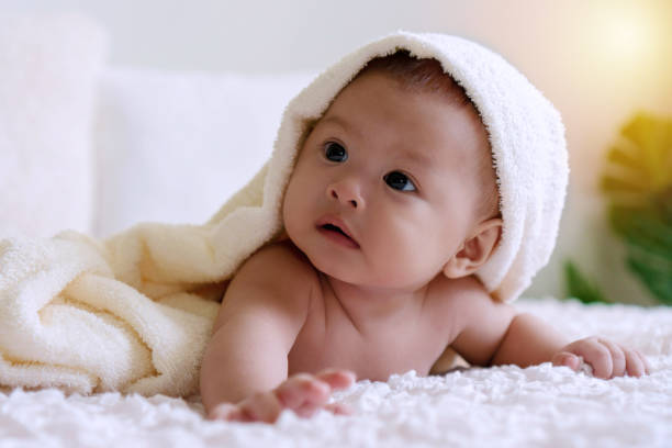 istock Cheerful cute baby lying under white blanket looking at something. Innocence baby crawling on white bed with towel on his head at home. 1302803504
