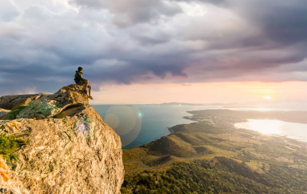 stunning view of a boy enjoying a beautiful sunset sitting on top of a mountain, golfo aranci, sardinia, italy. - climbing men sea cliff imagens e fotografias de stock