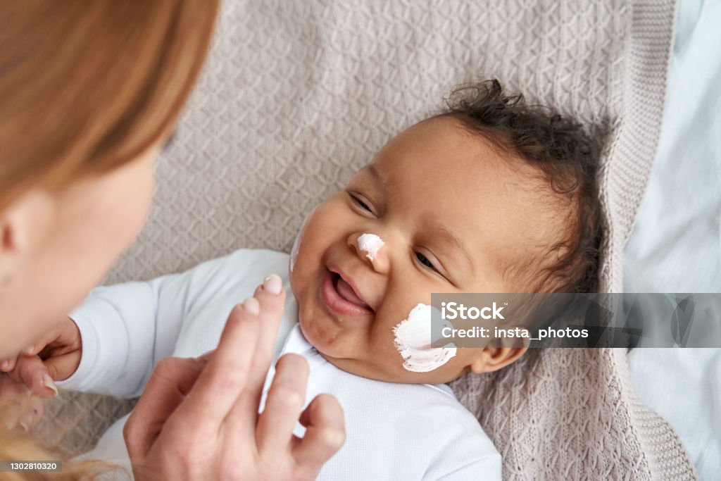 Caucasian mother applying healthy skin care moisturizing cream on cute adorable funny african american baby girl daughter face. Skincare lotion for infant pediatric diathesis treatment concept. Baby - Human Age Stock Photo
