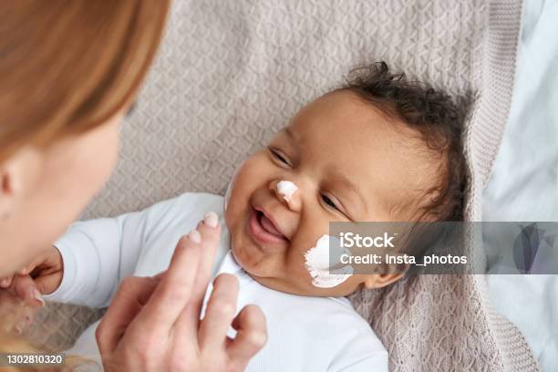Caucasian Mother Applying Healthy Skin Care Moisturizing Cream On Cute Adorable Funny African American Baby Girl Daughter Face Skincare Lotion For Infant Pediatric Diathesis Treatment Concept Stock Photo - Download Image Now