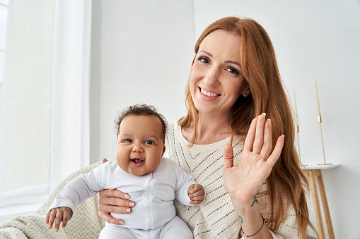 Happy young caucasian mother waving hand holding little cute funny african american baby girl daughter on lap having virtual distance meeting video call looking at camera at home, webcam view.
