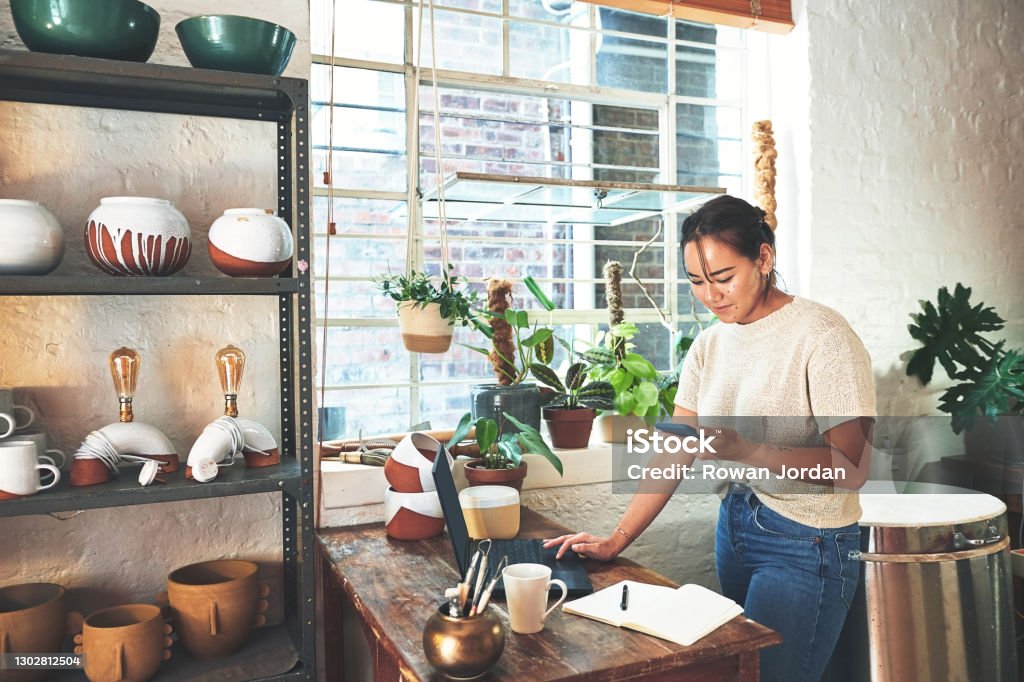 This customer wants something specific Cropped shot of an attractive young business owner standing and using her laptop and cellphone in her pottery studio Small Business Stock Photo
