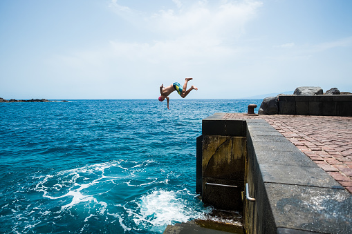 one young man or teenager enjoying the summer time and vacations outdoors at the beach jumping of a cliff to the water doing a front flip - active people having fun traveling
