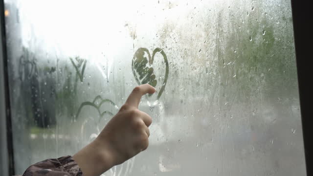 A child draws a heart with his finger on the fogged glass of a bus traveling through the city on a gloomy, rainy day. Cars are driving outside the window. The concept. Faceless. Close-up. 4K.