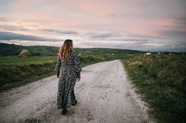 Rear view of a young woman wearing a dress walking on a dirt road in a rural area at sunset