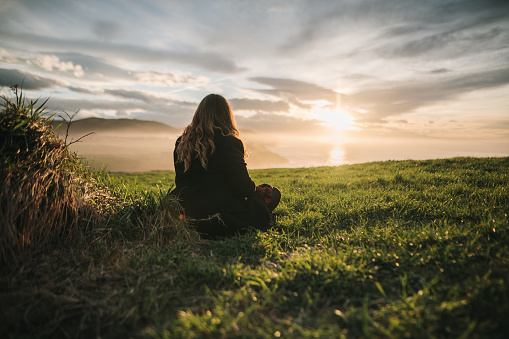 Young woman sitting on a green grass field by the sea at sunset