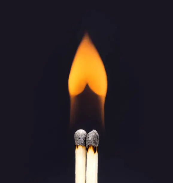 Photo of Heart shaped flame coming from a pair of burning matchsticks on a black background.