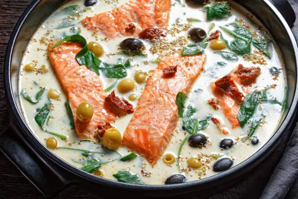 Tuscan salmon dish of pan-seared skin-on salmon fillet with cream sauce, garlic, leek, sun-dried tomatoes, olives, and spinach on a black  baking dish on a dark wooden table, top view, close-up