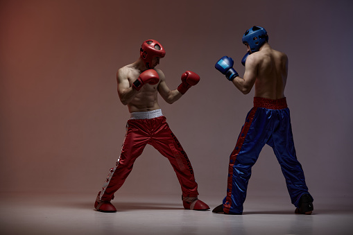 Wrestling of two fighting males in helmets and boxing gloves in red light in studio, martial arts, mixed fight concept. High quality photo
