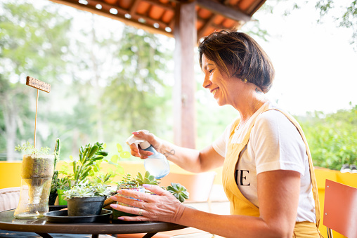 Smiling mature woman spraying water on some potted plants on a table on her patio at home