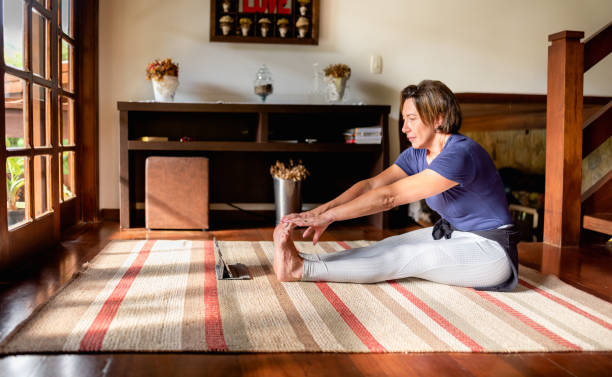 Woman stretching at home during a streaming yoga class on a tablet Mature woman touching her toes while doing yoga in her living room online with a digital tablet touching toes stock pictures, royalty-free photos & images
