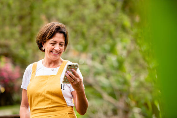 Smiling mature woman video calling on a phone outside in her yard Smiling mature woman video calling on a smart phone while standing outside in her back yard brazilian ethnicity stock pictures, royalty-free photos & images