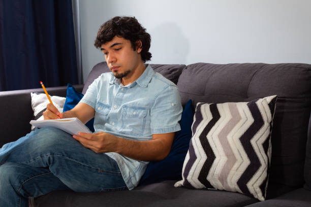 istock Young hispanic man with wavy hair studying from home writing in a notebook with a pencil, sitting on a sofa 1302853179