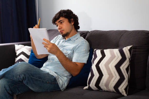 istock Young hispanic man with wavy hair studying from home writing in a notebook with a pencil, sitting on a sofa 1302853180