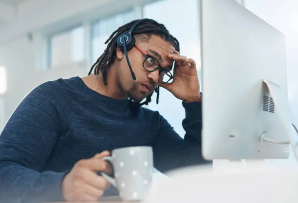 Shot of a young businessman wearing a headset and looking stressed out while working on a computer in an office