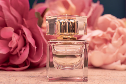 Glass bottle of fragrant eau de toilette against a background of pink peonies. Female perfume concept. Mock up. Close up photo
