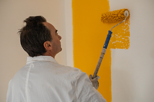 Photo from behind of mature man painting wall in yellow with paint roller.