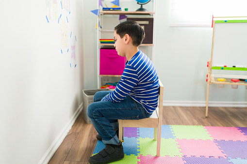Caucasian elementary boy sitting alone in a chair and facing the wall. Little boy grounded by his therapist or parents