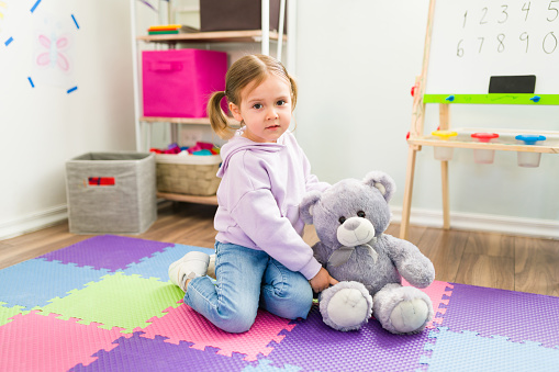 Portrait of an adorable elementary girl holding and playing with a teddy bear while sitting on a foam mat in the playroom