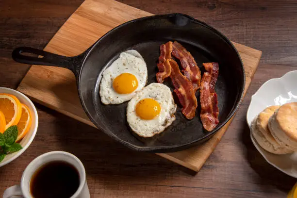 Bacon and eggs served in a cast iron skillet. The wooden table is full of other breakfast food items. Focus is on the bacon.