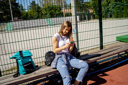 One beautiful young woman sitting on the bench and texting on mobile phone on basketball court on sunny day