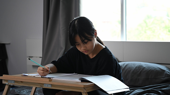 Asian children girl sitting on bed and doing homework.