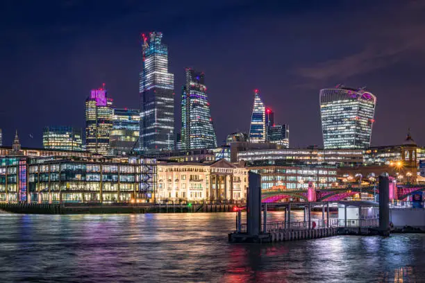 City of London's financial district and Tower of London famous landmark and tourist attraction during night hours with its evening illumination as photographed from Southbank of the river Thames. Long exposure technique used to blur the river Thames high tide waters reflecting the city lights and nautical transportation ferries. Shot on EOS R full frame system with premium RF lens for highest quality and high resolution. Blue toning added in post production.