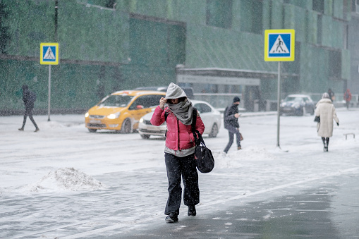 Moscow. Russia. February 12, 2021. City street, passer-by and snowfall. A woman walks down the street in winter and covers her face with a hood from the wind, snow is falling. Selective focus.