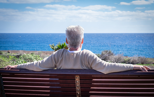 Rear view of an aged white-haired man relaxing in front to the sea, sitting on a bench looking at the horizon. Senior man enjoying retirement
