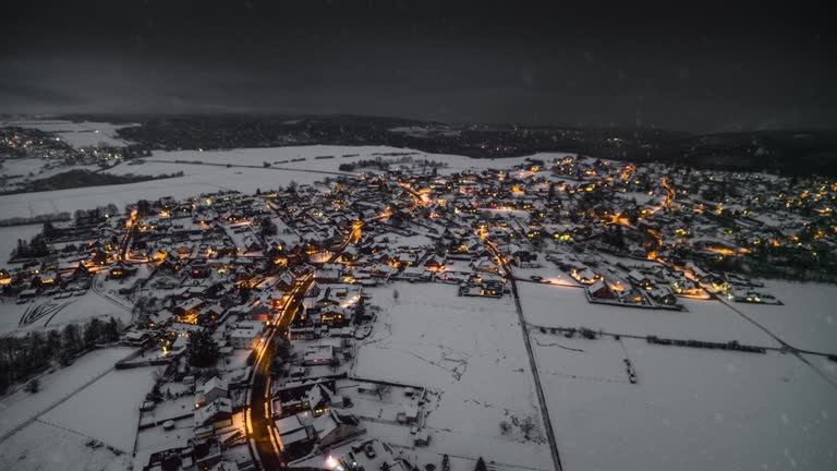 Aerial shot of a small german village in a snow covered winter landscape. Snow flakes are falling from the sky. The warm illumination from the street lights and houses are forming a beautiful contrast to the cold snow covered surrounding. Eifel mountain range in Germany - Europe.