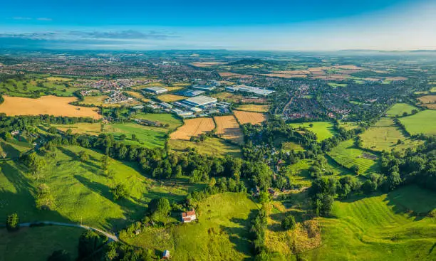 Suburban homes, gardens and factories surrounded by patchwork quilt landscape of green pasture, golden crops and wooded hills in this aerial panoramic vista.