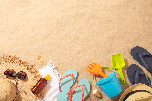Top view of a beach towel with a sun straw hat, a sun tan and a sunglasses on top surrounded by a flip flops, sand bucket and some sea shells. All the objects are at the lower side of the image leaving a useful copy space at the top on a beach sand background. Predominant colors are brown, navy and Light blue. Studio shot taken with Canon EOS 6D Mark II and Canon EF 24-105 mm f/4L