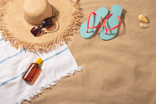 Top view of a beach towel with a sun straw hat, a sun tan and a women sunglasses on top surrounded by a flip flops and some sea shells. All the objects are at the top and at the left side of the image leaving a useful copy space at the lower right corner on a beach sand background. Predominant colors are brown and Light blue. Studio shot taken with Canon EOS 6D Mark II and Canon EF 24-105 mm f/4L