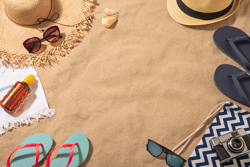 Top view of a beach towel with a sun straw hat, a sun tan, a vintage camera and a sunglasses on top surrounded by a flip flops and some sea shells. All the objects are at the borders of the image leaving a useful copy space at the center on a beach sand background. Predominant colors are brown, navy and Light blue. Studio shot taken with Canon EOS 6D Mark II and Canon EF 24-105 mm f/4L