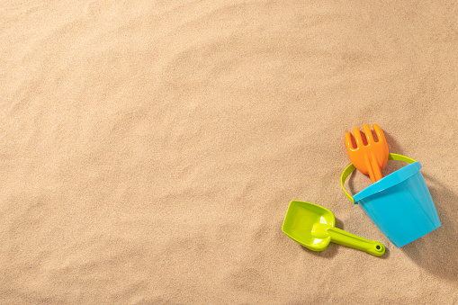 Top view of a blue sand bucket with a green pale and a orange shovel on a beach sand background. All the objects are at the lower left corner leaving a useful copy space at the left and at the top of the image. Studio shot taken with Canon EOS 6D Mark II and Canon EF 24-105 mm f/4L