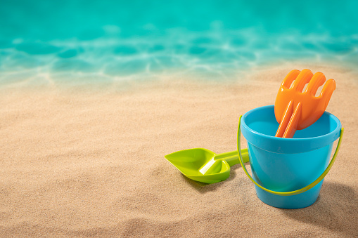 Front view of a blue sand bucket with a green pale and an orange shovel laying at the lower right corner of the image leaving a useful copy space at the top and at the left side on a beach background. Studio shot taken with Canon EOS 6D Mark II and Canon EF 24-105 mm f/4L
