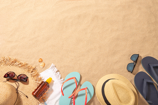 Top view of a beach towel with a sun straw hat, a sun tan, a vintage camera and a sunglasses on top surrounded by a flip flops and some sea shells. All the objects are at the lower side of the image leaving a useful copy space at the top on a beach sand background. Predominant colors are brown, navy and Light blue. Studio shot taken with Canon EOS 6D Mark II and Canon EF 24-105 mm f/4L