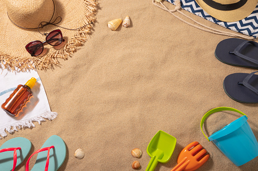 Top view of a beach towel with a sun straw hat, a sun tan and a sunglasses on top surrounded by a flip flops, sand bucket and some sea shells. All the objects are at the borders of the image leaving a useful copy space at the center on a beach sand background. Predominant colors are brown, navy and Light blue. Studio shot taken with Canon EOS 6D Mark II and Canon EF 24-105 mm f/4L