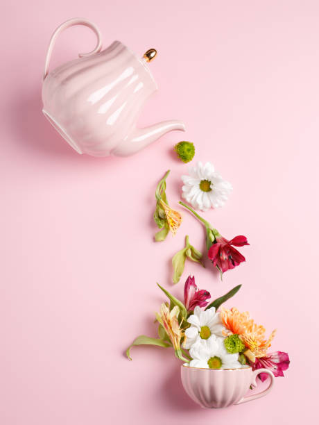 Creative layout with tea pot pouring fresh flowers and leaves into tea cup on pastel pink background