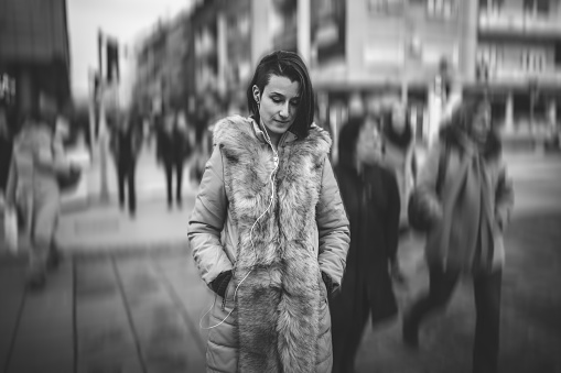Sad young woman in fur jacket listening music and walking downtown.