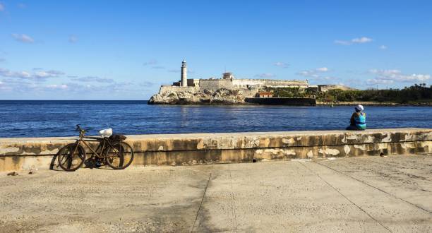 Malecon Promenade and Morro Castle in Havana Havana, Cuba, November 20, 2017: A woman is sitting on the wall of Malecon Promenade and looking on the Castillo De Los Tres Reyes Del Morro (Morro Castle) - a fortress guarding the entrance to Havana bay in Havana. morro castle havana stock pictures, royalty-free photos & images