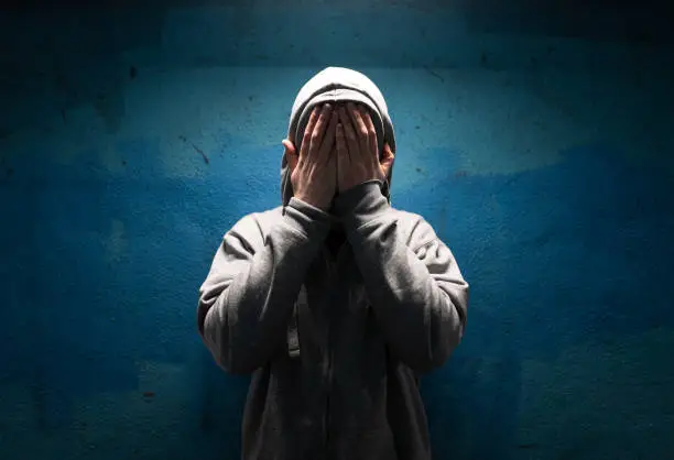 Color image depicting an adult man wearing a grey hoodie with the hood over his head in an underground subway tunnel. His expression is one of despair, sadness and depression. His hands are covering his face in a gesture of bleak desperation and hopelessness. Room for copy space.