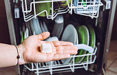 Selective focus on woman hand, holding homemade natural dishwasher pod defocus dishes in dishwasher on background. Green sustainable lifestyle concept.