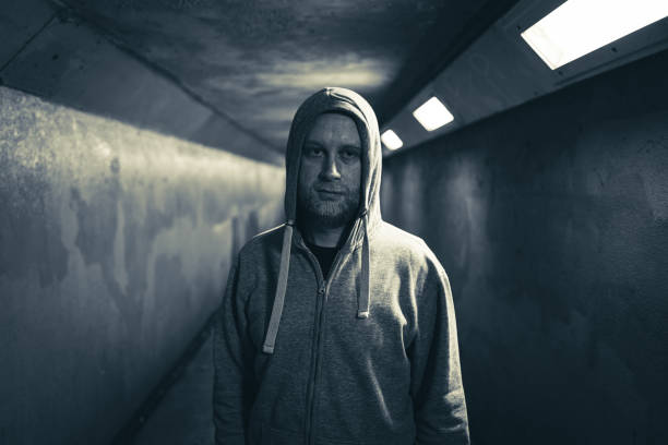 Color image depicting an adult man in his 30s in an underground subway tunnel. He is wearing a hooded sweater with the hood pulled up over his head. His expression is one of despair, sadness and depression. Focus is on the man in the foreground while we can see the light at the end of the dark and moody tunnel in the background. Room for copy space.