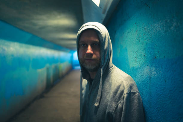 Color image depicting an adult man in his 30s in an underground subway tunnel. He is wearing a hooded sweater with the hood pulled up over his head. His expression is one of despair, sadness and depression. Focus is on the man in the foreground while we can see the light at the end of the dark and moody tunnel in the background. Room for copy space.