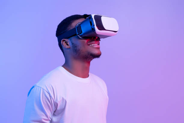 Modern technologies for entertainment. Excited black man wearing vr glasses looking aside Modern technologies for entertainment. Excited black man wearing vr glasses looking aside at copy space, african guy experiencing virtual reality, standing on purple background in neon lighting virtual reality simulator stock pictures, royalty-free photos & images